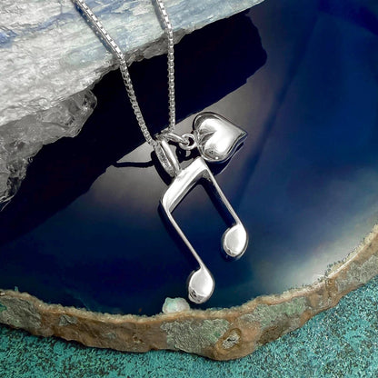 A silver necklace with a heart-shaped pendant and a musical note pendant is displayed on a blue and green textured surface.