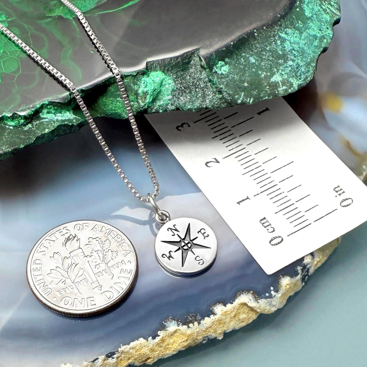 a silver necklace with a compass pendant, a United States quarter, and a ruler on a green and gray rock.
