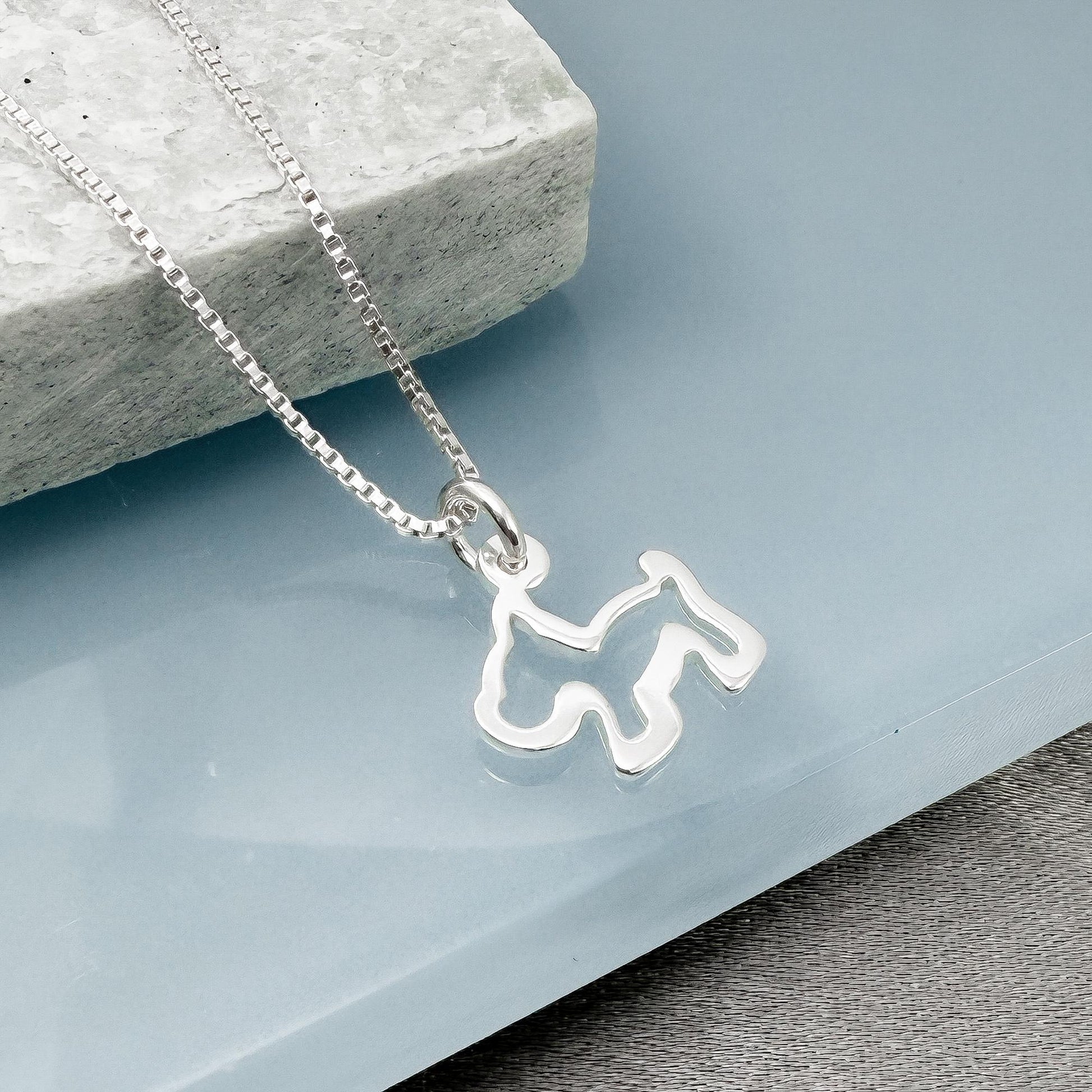 A silver necklace with a pendant in the shape of a dog silhouette is displayed on a light blue surface.