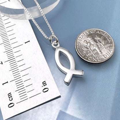 A silver fish pendant necklace and a United States one dollar coin are placed on a blue surface, with a ruler for scale.
