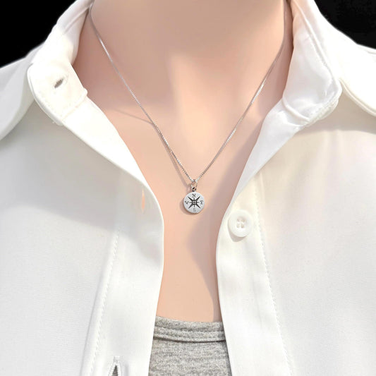 A silver necklace with a round pendant is worn around a person's neck, who is wearing a white collared shirt.