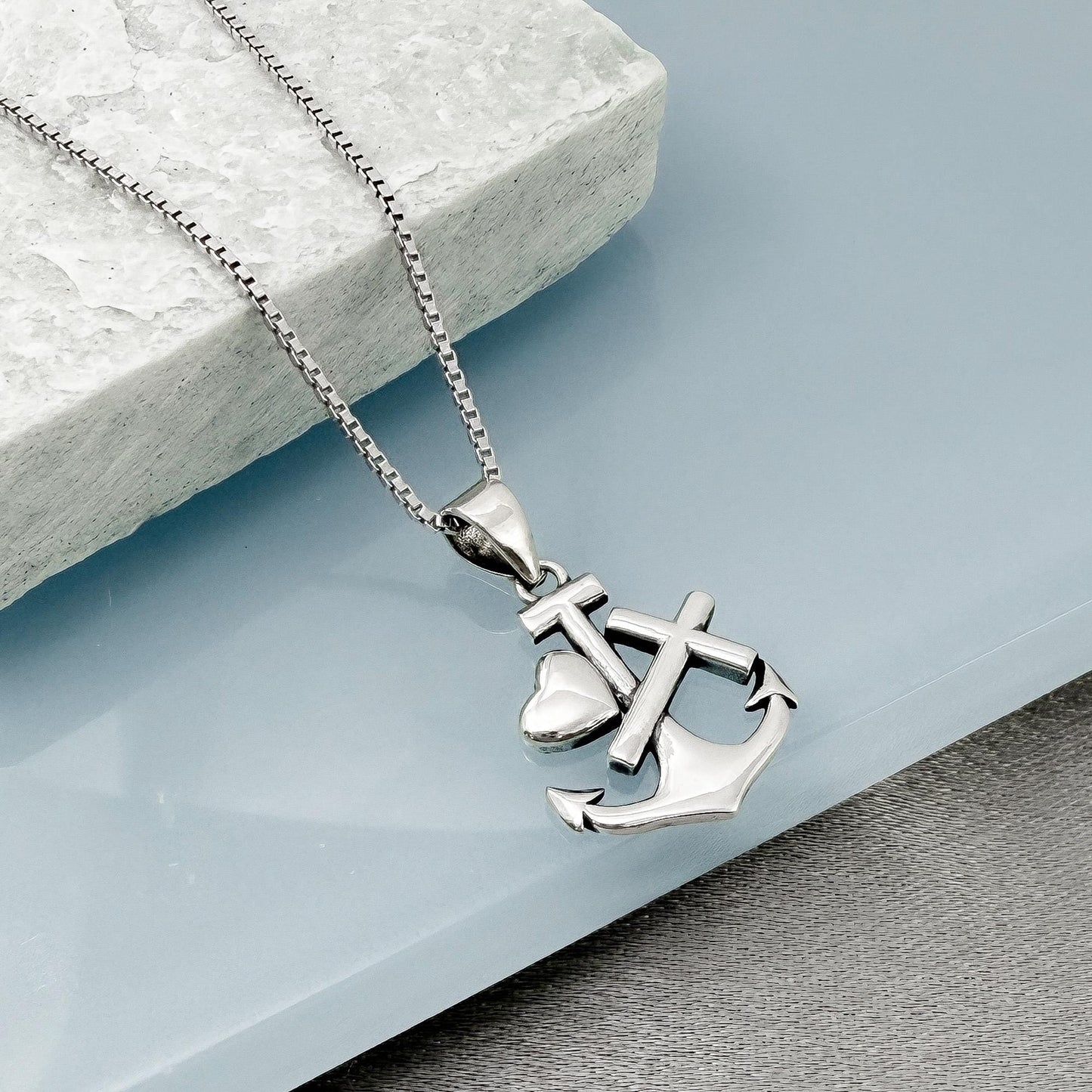 A silver-toned necklace with a pendant featuring an anchor and a heart symbol, resting on a gray surface.