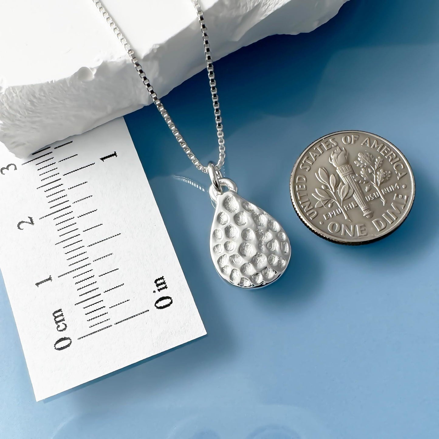 a silver necklace with a pendant, a silver coin, and a white paper with measurements on it, all placed on a blue surface.