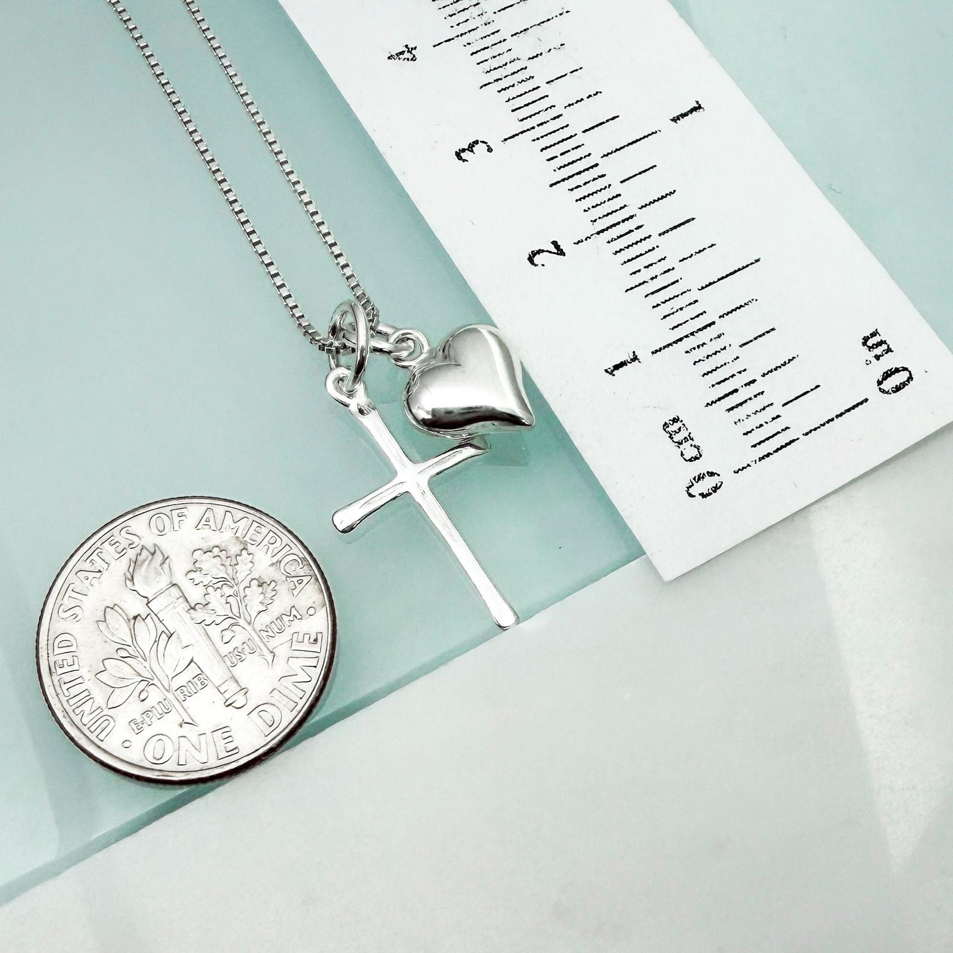 a silver cross pendant and a silver heart charm, both of which are part of a necklace. The necklace is placed next to a United States quarter coin, which is approximately 1 inch in diameter.