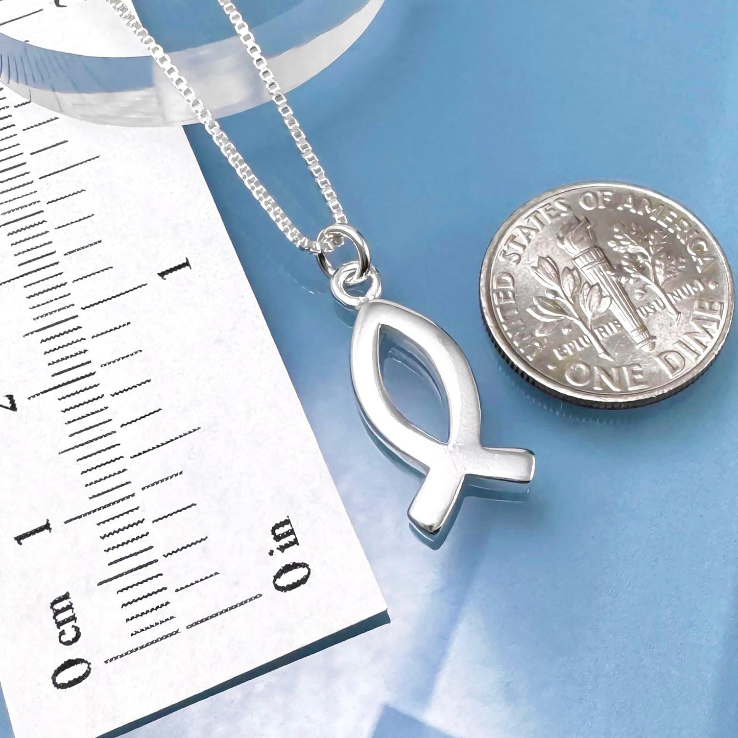 A silver fish pendant necklace and a United States one dollar coin are placed on a blue surface, with a ruler for scale.