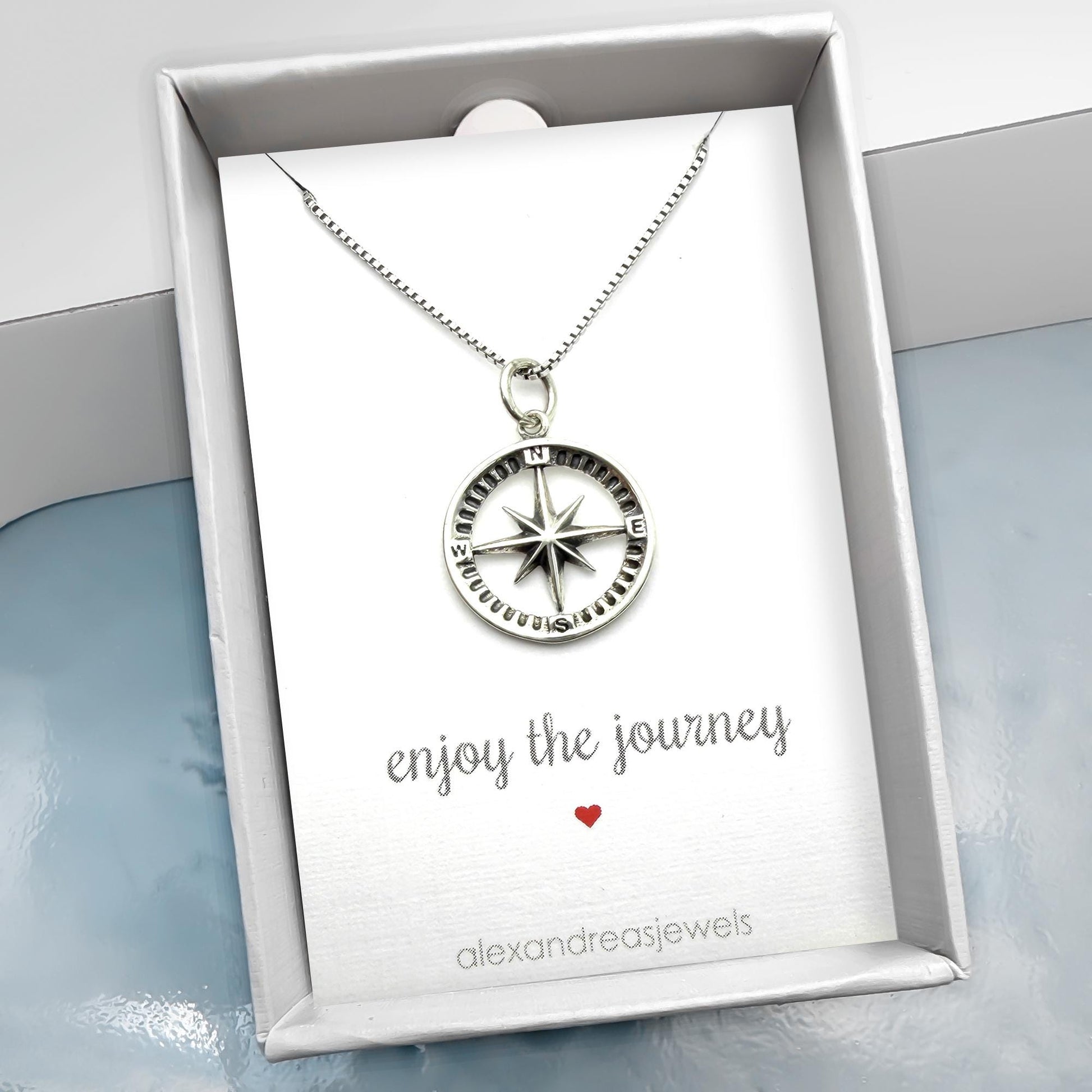 A silver compass pendant necklace is displayed in a white box with a message that reads "Enjoy the journey" and a red heart symbol.