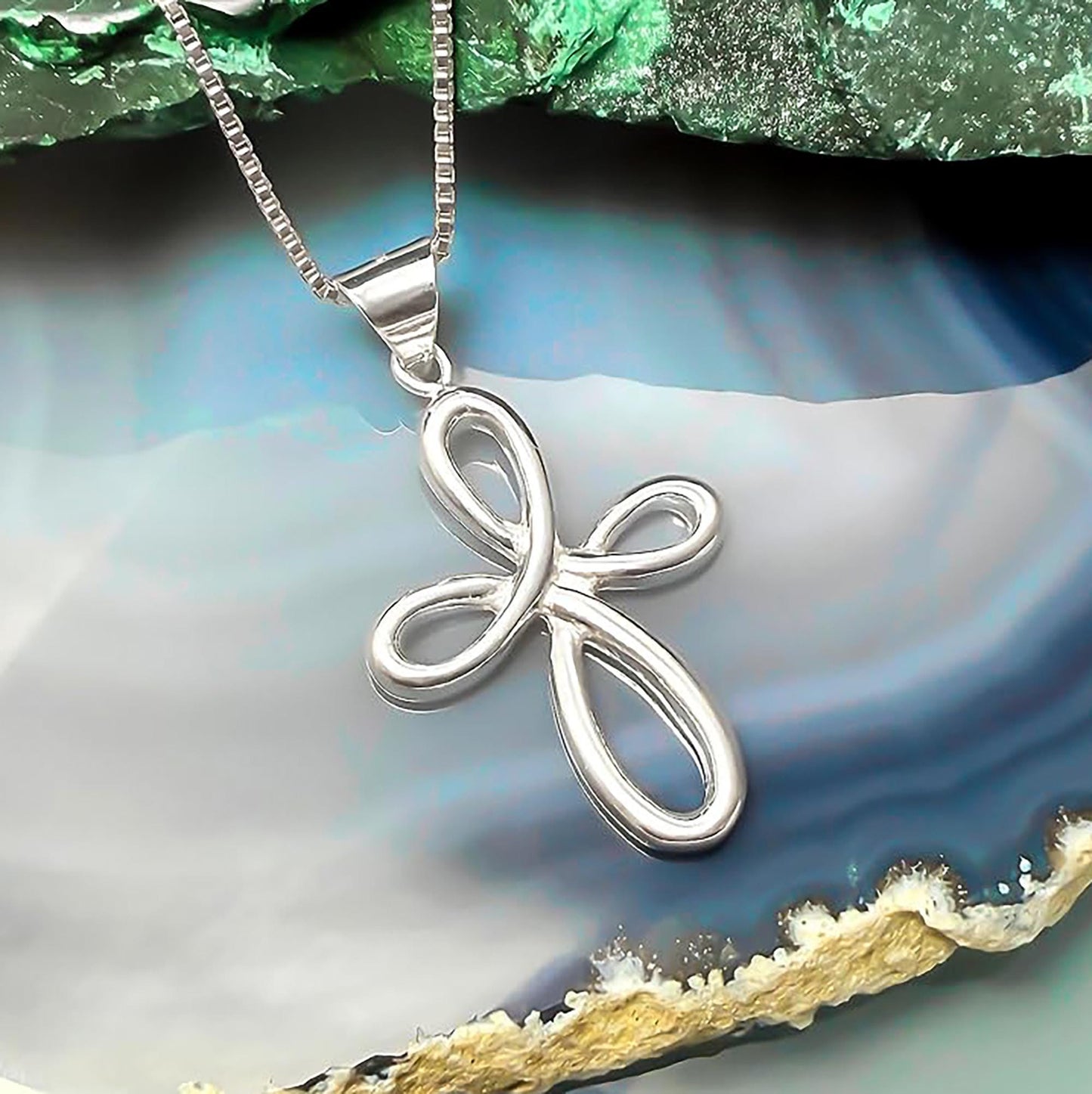 A silver cross pendant with a twisted design is displayed on a chain against a blue and green background.
