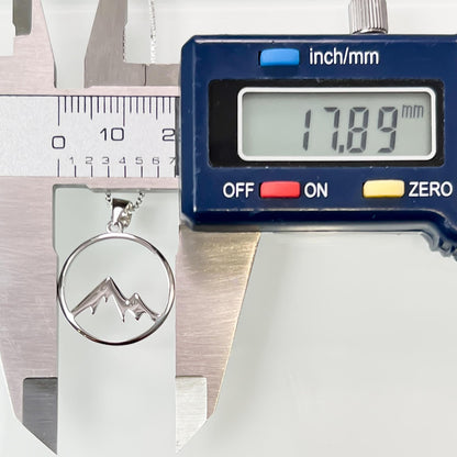 A silver pendant necklace with a mountain symbol is being measured against a blue digital caliper, which displays the size in millimeters.