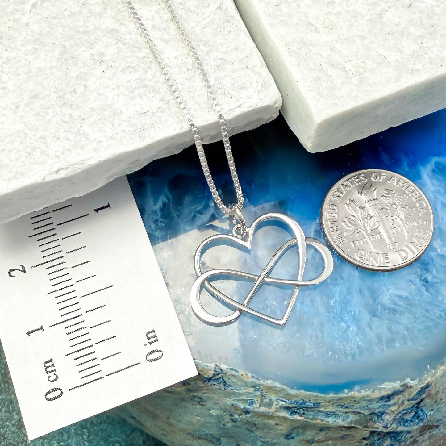 a silver heart-shaped pendant necklace with a silver chain, placed on a blue and white marble surface. Next to the necklace is a silver coin, and there is a ruler for scale.