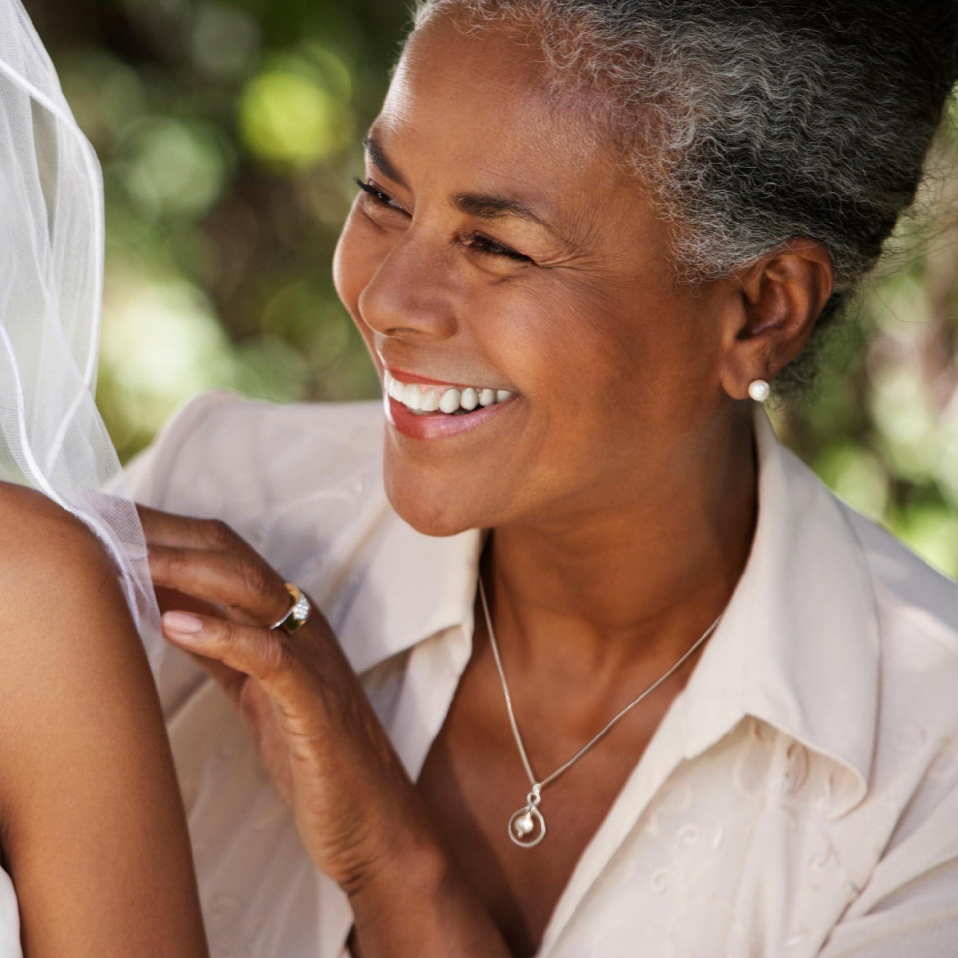 A smiling woman with gray hair wearing a white blouse and a necklace is shown in a close-up shot.