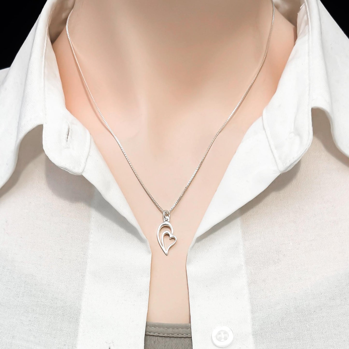 A silver necklace with a heart-shaped pendant is worn around a person's neck, who is wearing a white collared shirt.