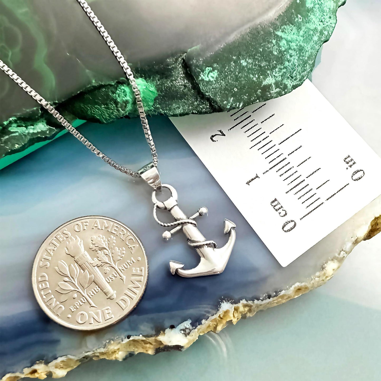 a silver anchor pendant hanging from a chain, placed next to a United States one-dollar coin.