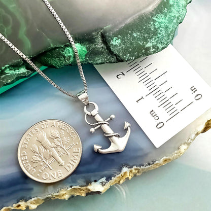 a silver anchor pendant hanging from a chain, placed next to a United States one-dollar coin.