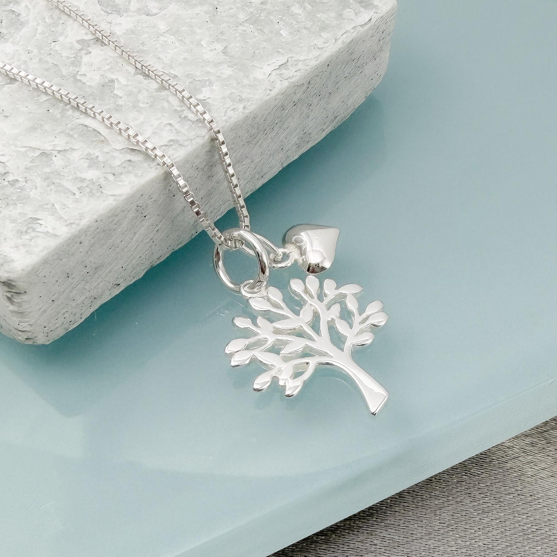 A silver necklace with a pendant featuring a tree design is displayed on a light blue surface.
