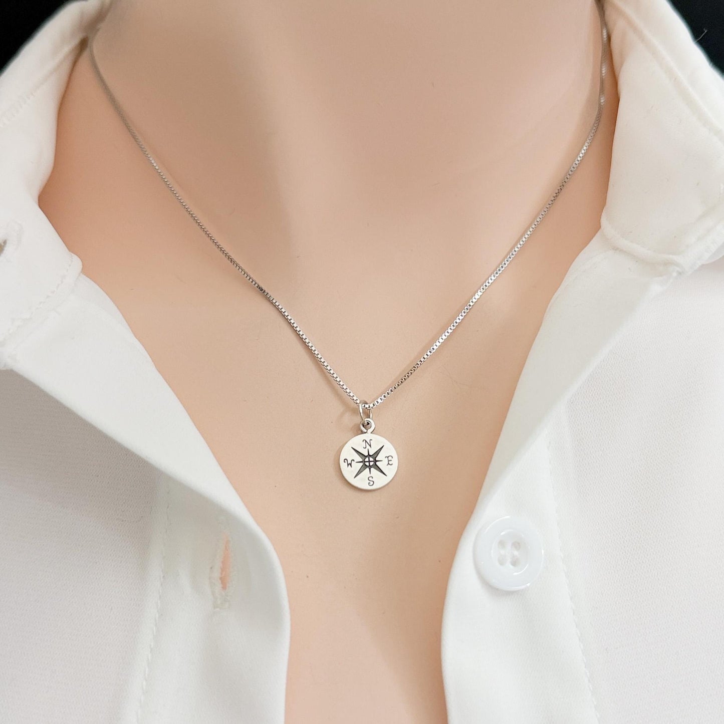 A silver necklace with a compass pendant is shown on a person wearing a white shirt.
