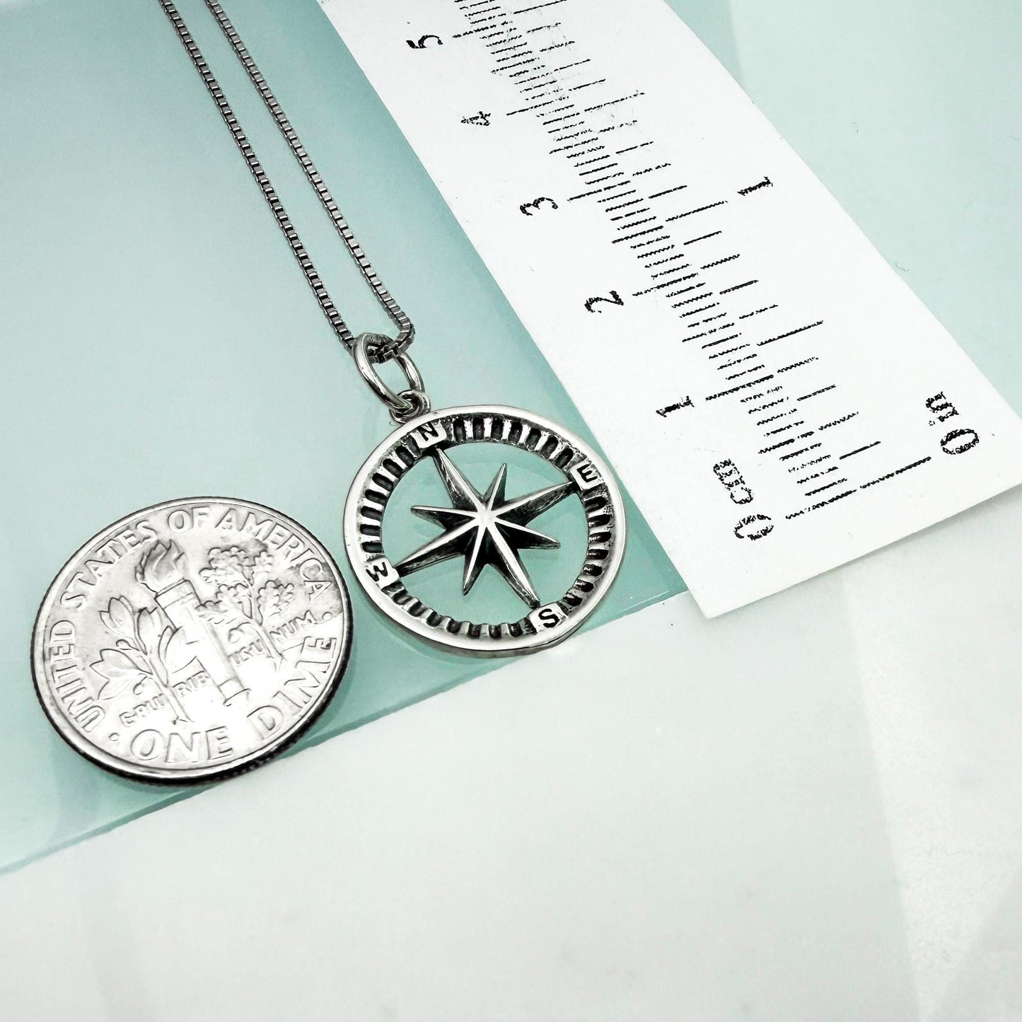 a silver compass pendant and a United States one-dollar coin, both placed on a light blue surface.