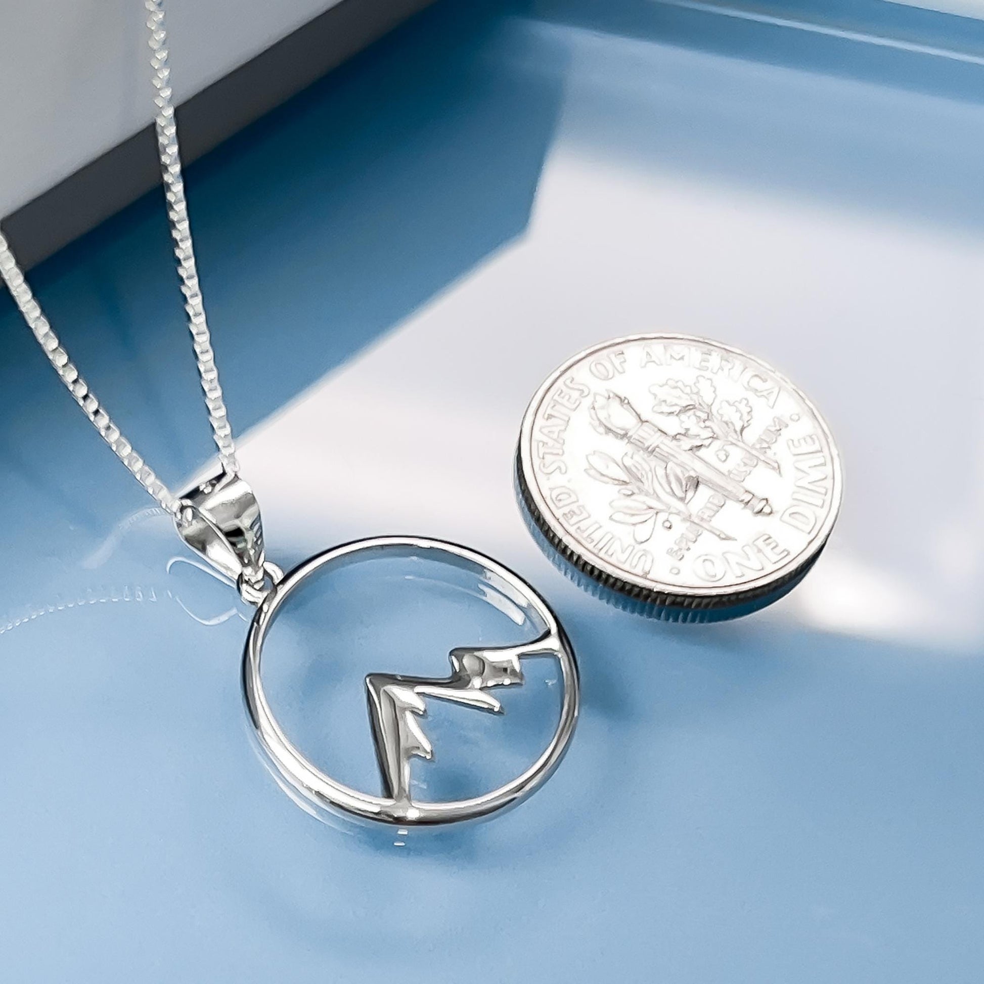 A silver necklace with a pendant in the shape of a mountain is placed next to a United States quarter on a blue surface.