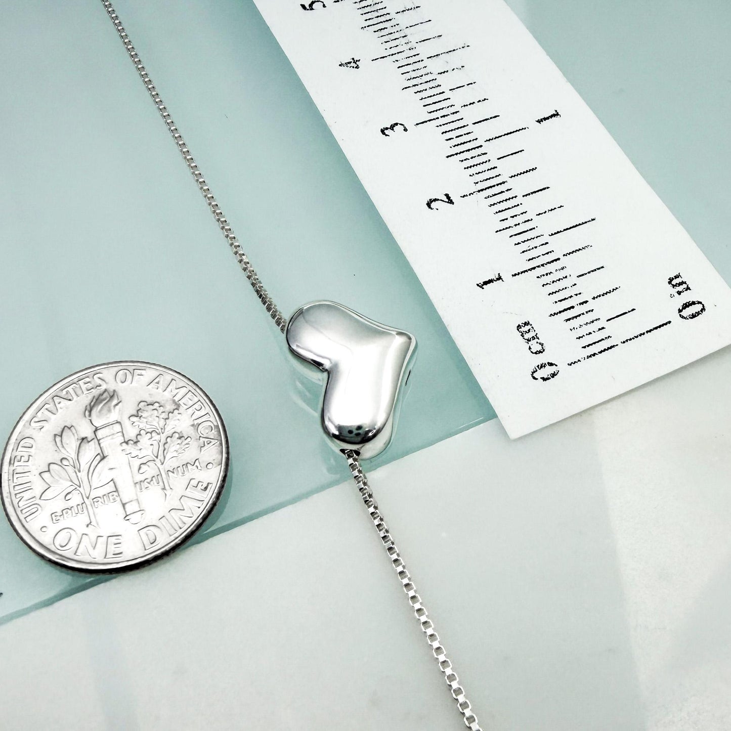 a silver heart-shaped charm attached to a chain, with a United States one-dollar coin placed next to it.