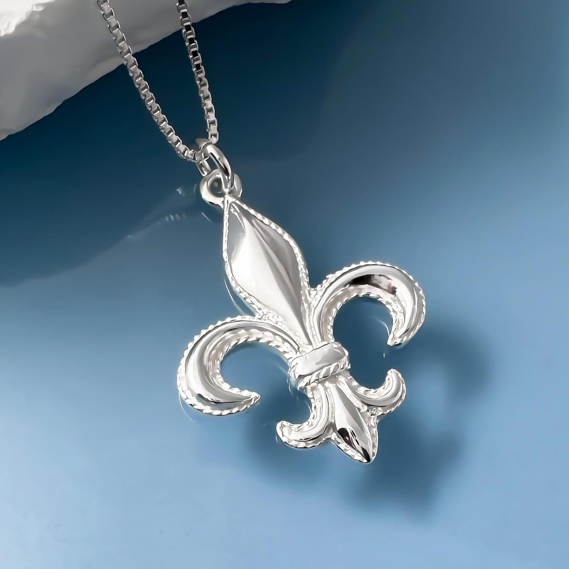 A silver-colored pendant in the shape of a fleur-de-lis, a heraldic symbol, is hanging from a chain against a blue background.