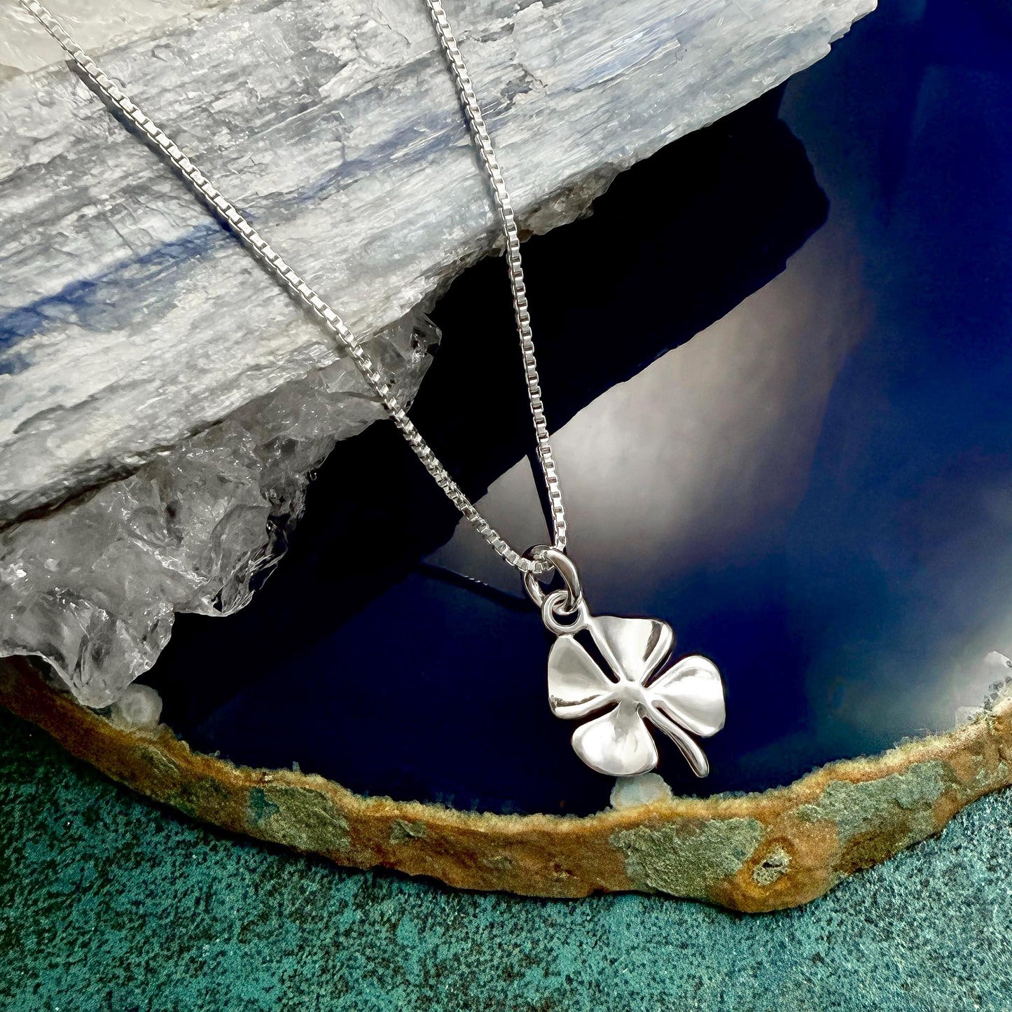 A silver cloverleaf pendant necklace is displayed on a blue and green textured surface, with a clear crystal or gemstone in the background.