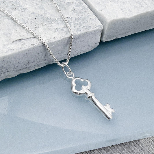 A silver-colored key pendant is hanging from a chain, resting on a white surface.