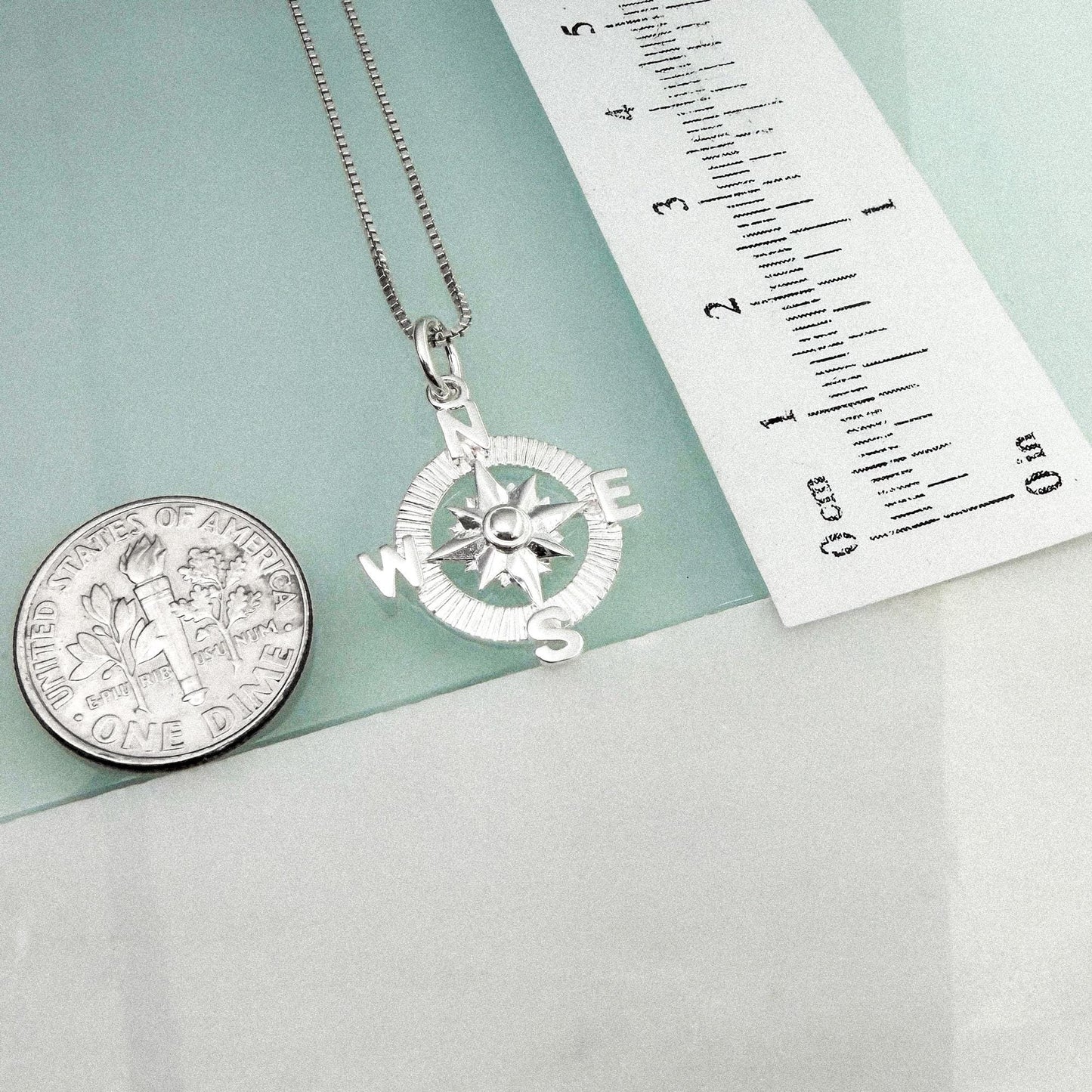 a silver compass pendant hanging from a chain, accompanied by a United States quarter and a ruler for size comparison.
