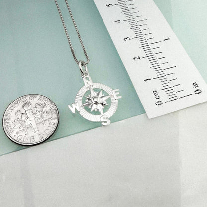 a silver compass pendant hanging from a chain, accompanied by a United States quarter and a ruler for size comparison.