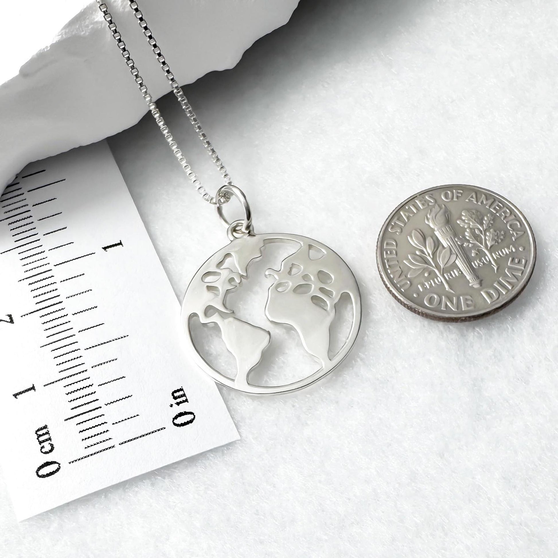 A silver necklace with a pendant in the shape of a world map, and a United States one dollar coin are placed next to each other on a white surface.
