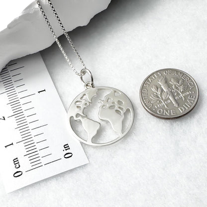 A silver necklace with a pendant in the shape of a world map, and a United States one dollar coin are placed next to each other on a white surface.