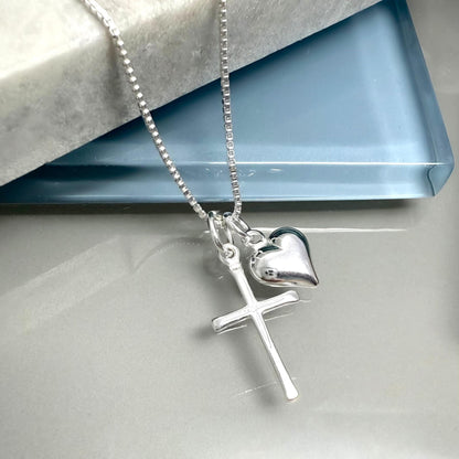 A silver cross necklace with a heart charm is displayed on a white surface.