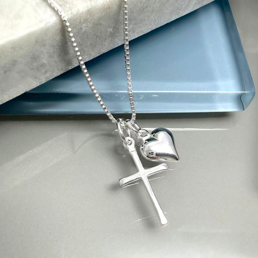 A silver cross necklace with a heart charm is displayed on a white surface.