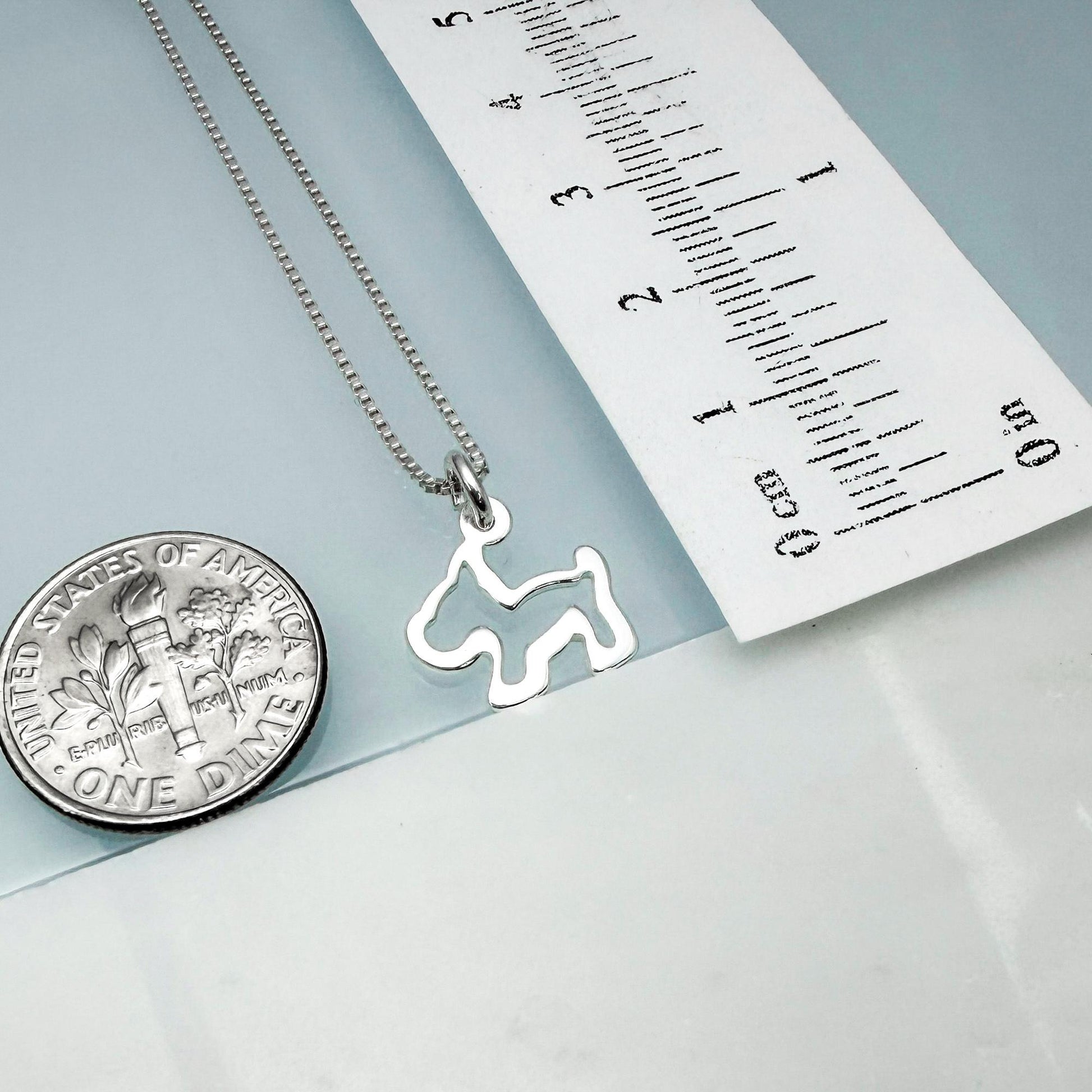 a silver dog-shaped pendant hanging from a chain, placed next to a United States one-dollar coin for scale.