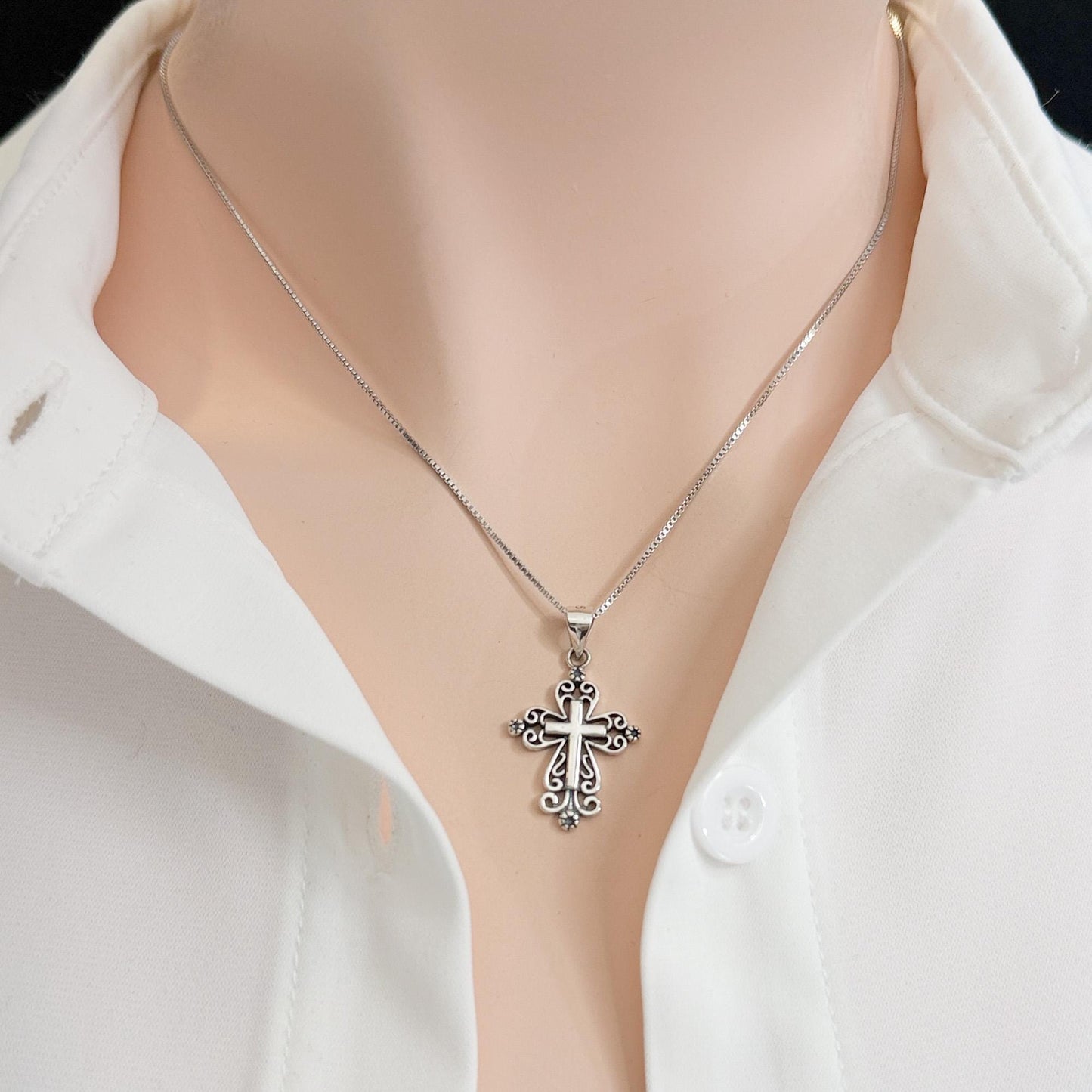 A silver cross pendant necklace is worn around a person's neck, with a white collared shirt visible underneath.