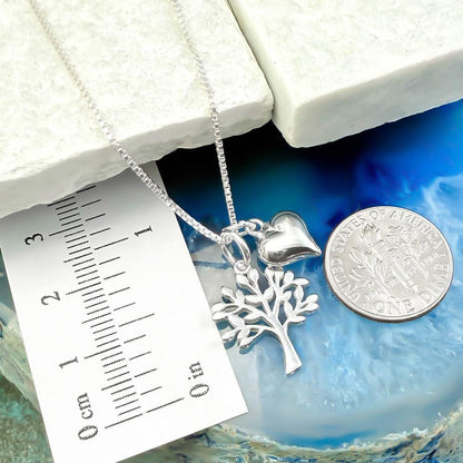 A silver necklace with a heart-shaped charm and a tree-shaped charm is displayed next to a U.S. quarter coin.