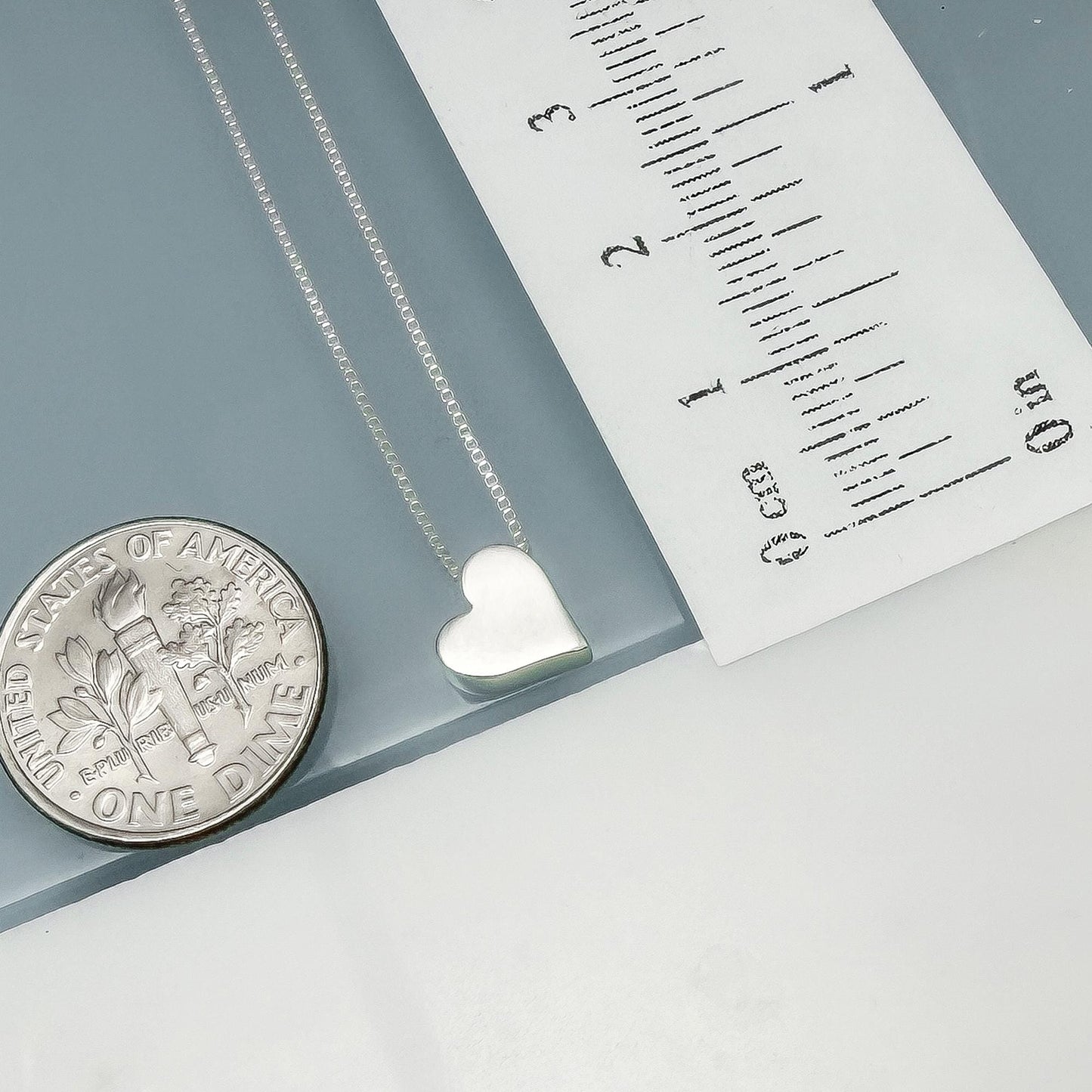 a silver heart-shaped pendant necklace and a United States one-dollar coin, with a ruler placed next to them.