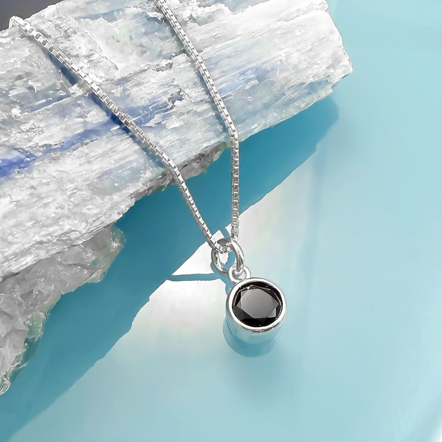 A silver necklace with a round black gemstone pendant is displayed against a blue background.
