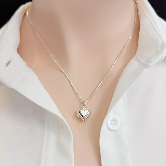 A silver heart-shaped pendant necklace is worn around a person's neck, with a white collared shirt visible underneath.