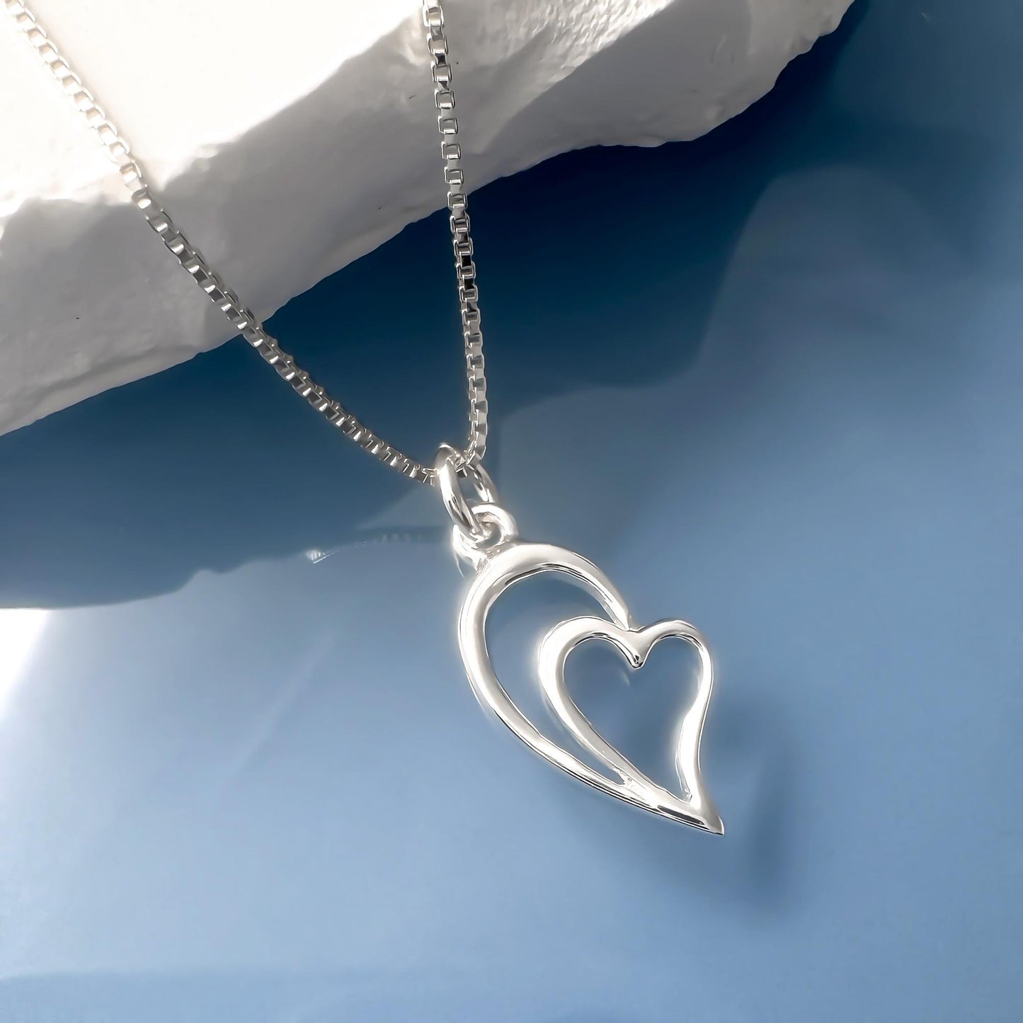 A silver heart-shaped pendant necklace with a chain is displayed against a blue background.