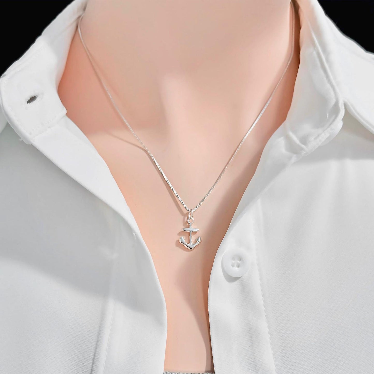 A silver necklace with a small anchor pendant is worn around a person's neck, who is wearing a white collared shirt.
