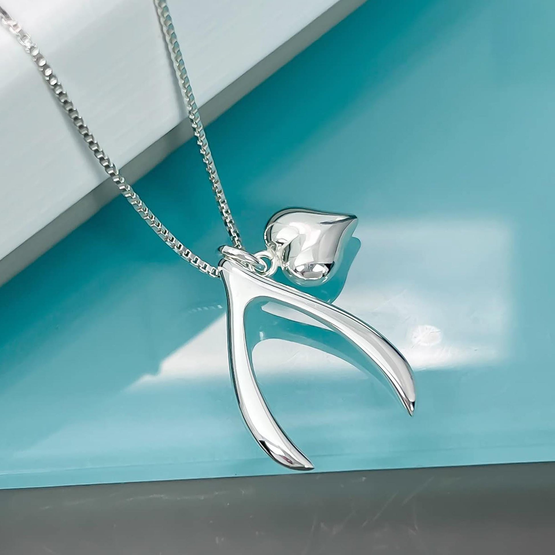 A silver necklace with a heart-shaped pendant and a silver-colored pendant is displayed against a turquoise background.