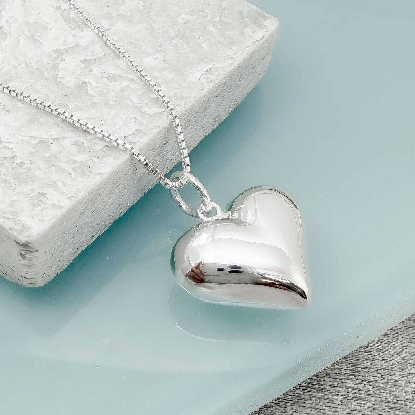A silver heart-shaped pendant necklace is displayed on a white stone surface.