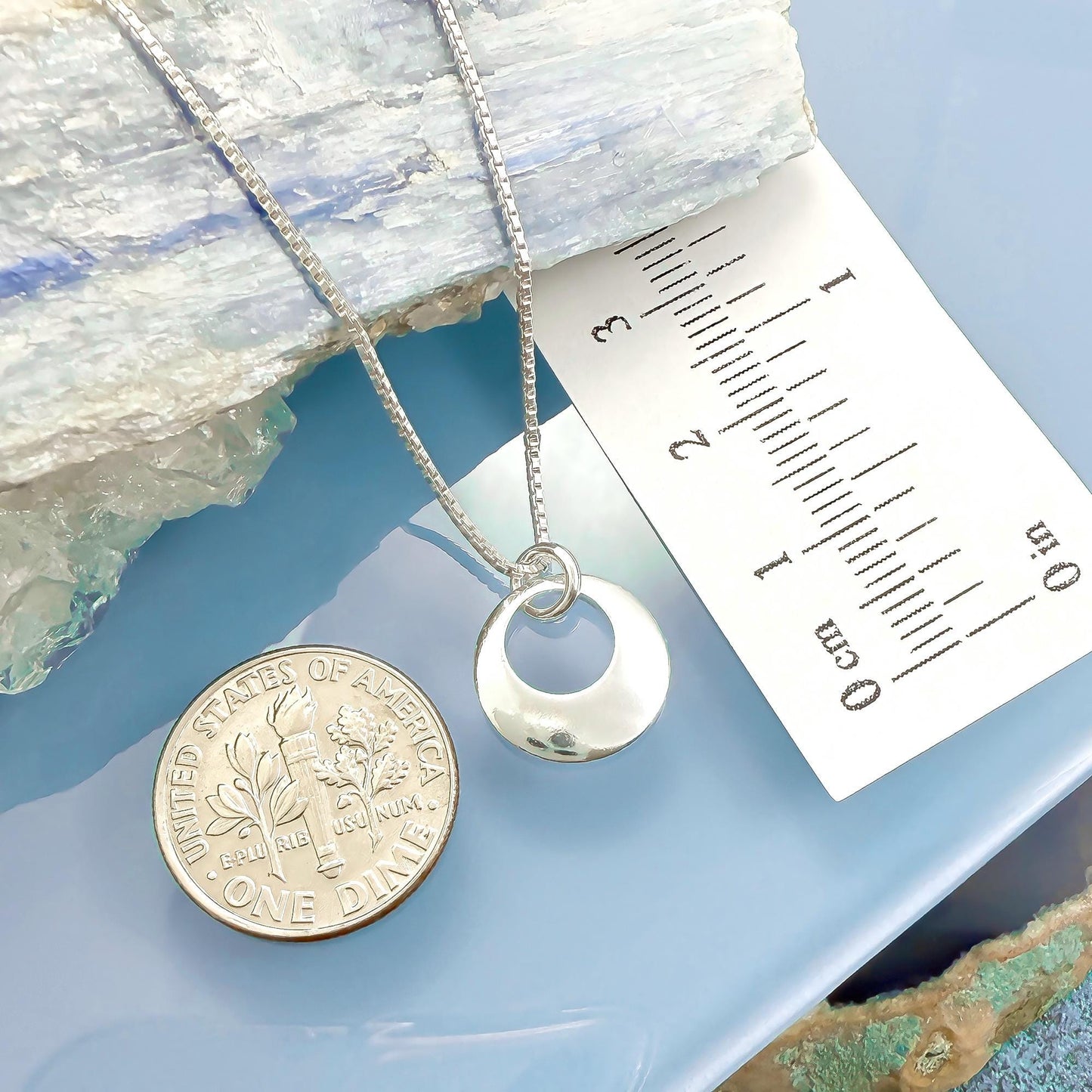 a silver necklace with a pendant, a silver coin, and a ruler on a blue surface.