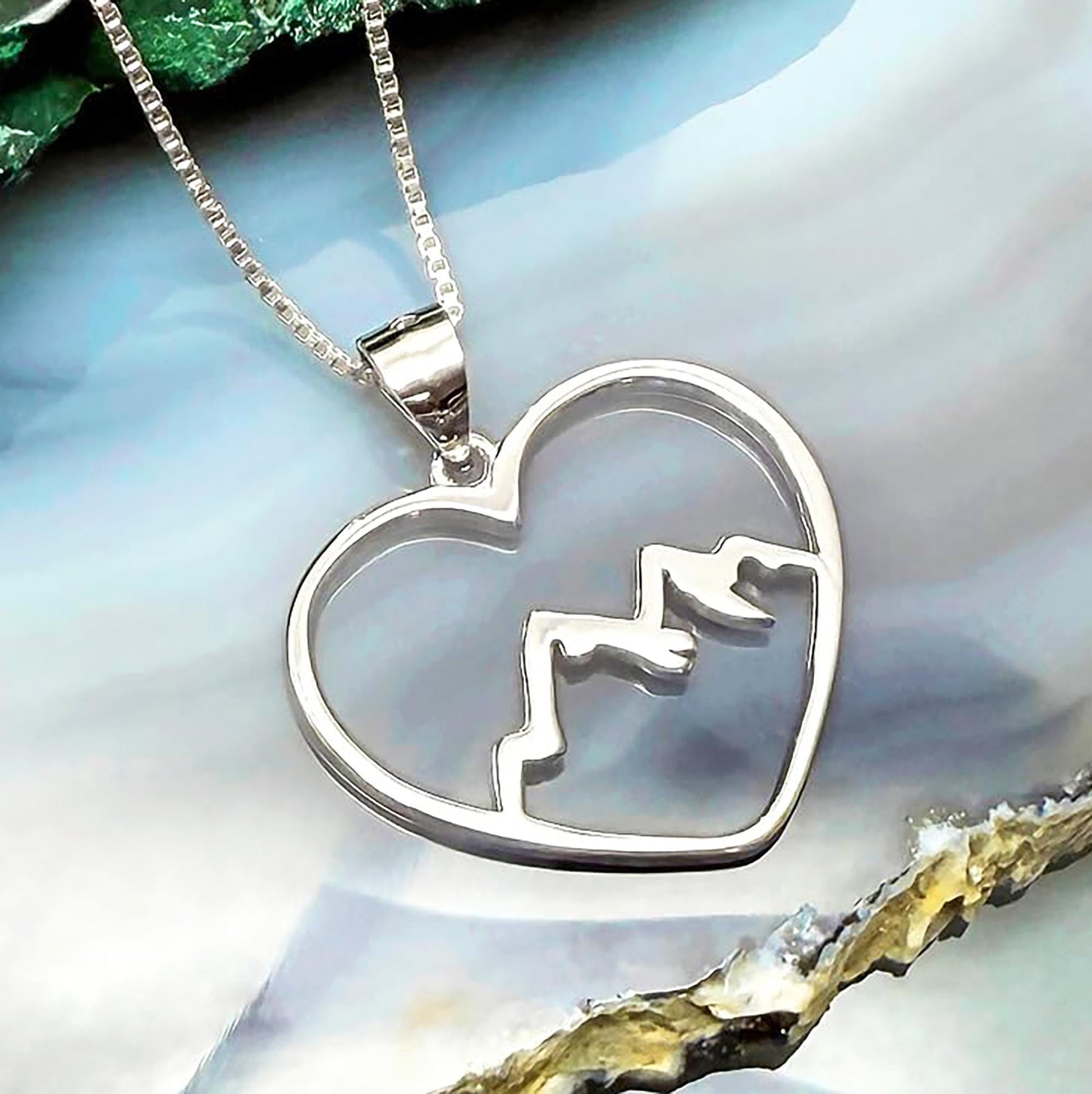 A silver heart-shaped pendant with mountain silhouettes is displayed against a blue and white background.
