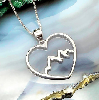 A silver heart-shaped pendant with mountain silhouettes is displayed against a blue and white background.