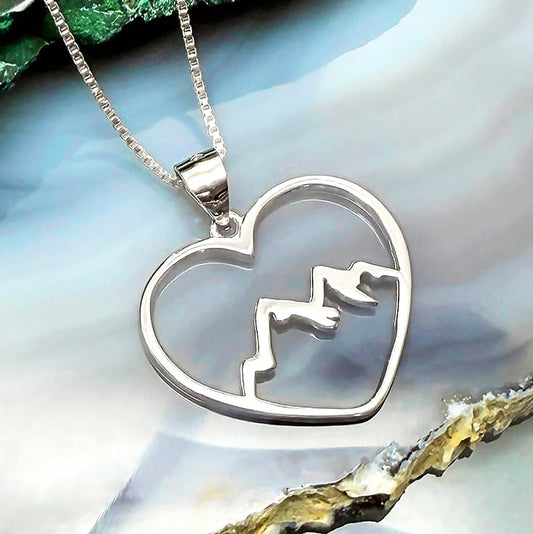 A silver heart-shaped pendant with mountain silhouettes is displayed against a blue and white background.