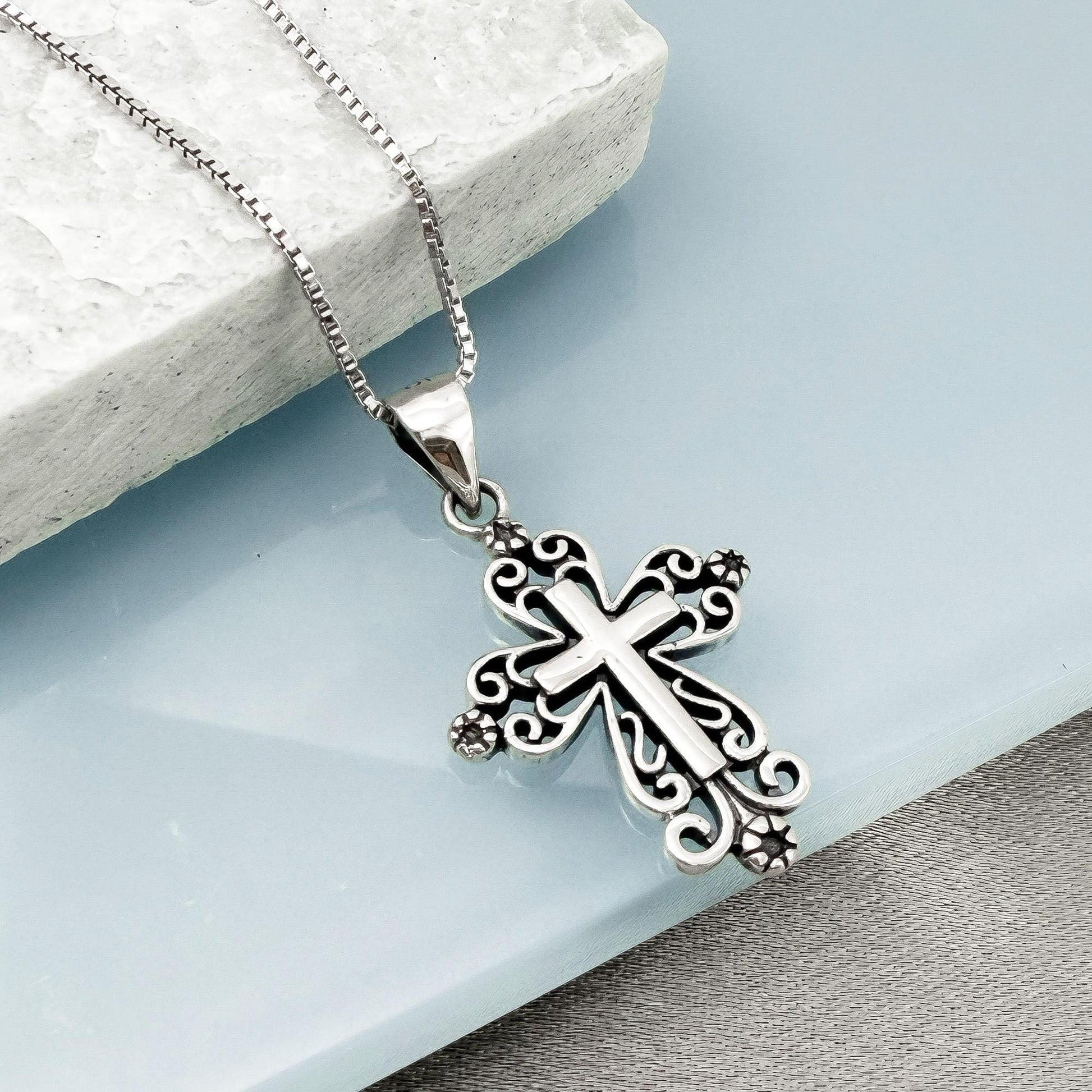 A silver cross pendant with intricate designs hangs from a chain against a light blue background.