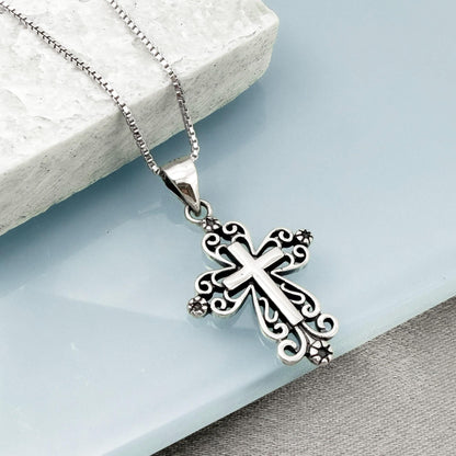 A silver cross pendant with intricate designs hangs from a chain against a light blue background.