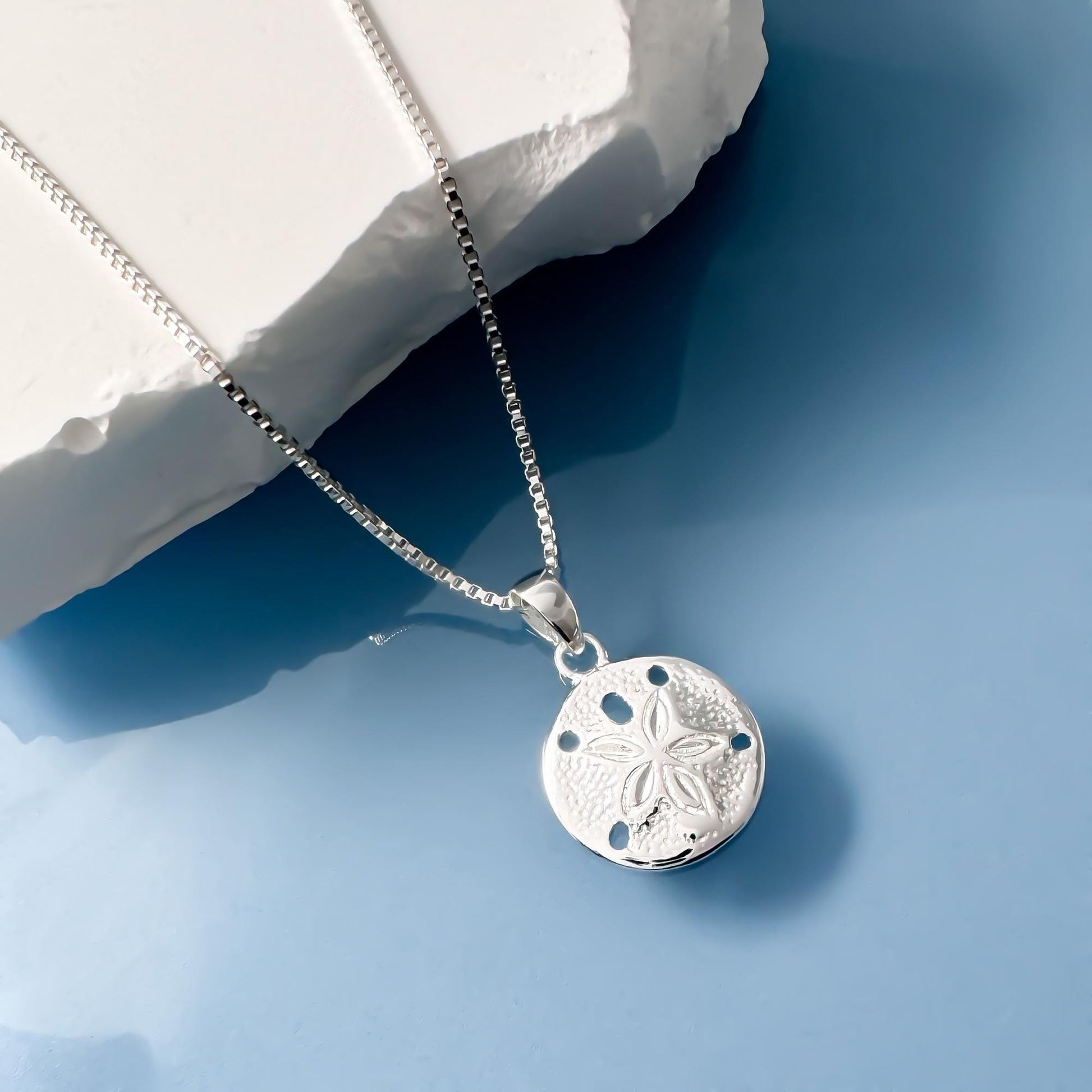 A silver necklace with a pendant featuring a flower design is displayed against a blue background.