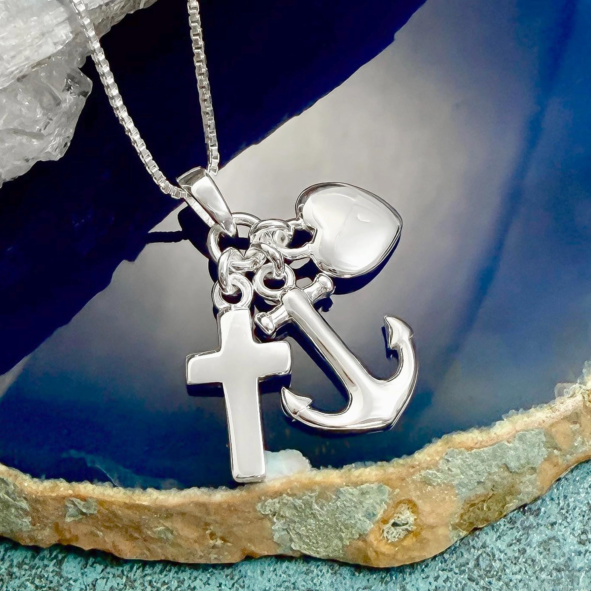 A silver necklace with a cross and an anchor pendant is displayed on a blue and white textured surface.