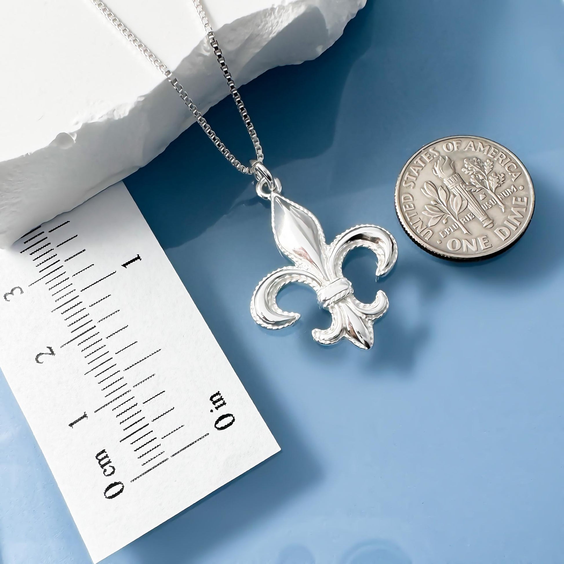 a silver fleur-de-lis pendant necklace, a silver coin, and a ruler on a blue surface.